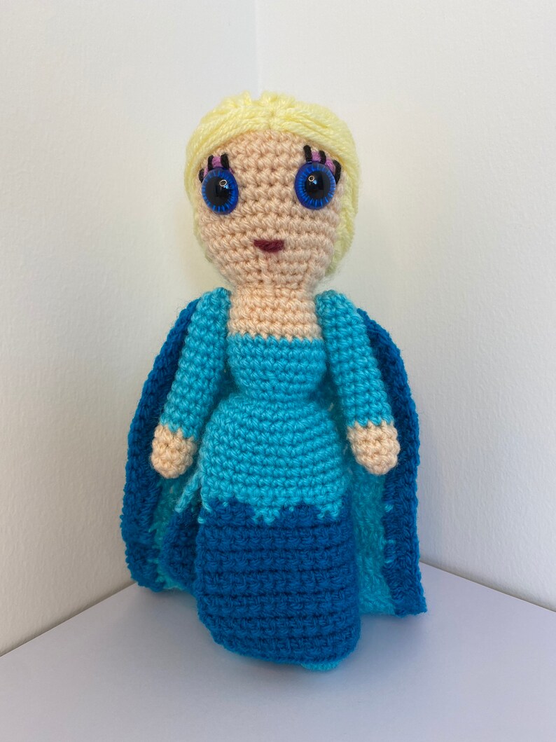 K&ouml;nnte beinhalten: Eine geh&auml;kelte Puppe mit blonden Haaren, blauen Augen und einem blauen Kleid. Die Puppe tr&auml;gt einen blauen Umhang.