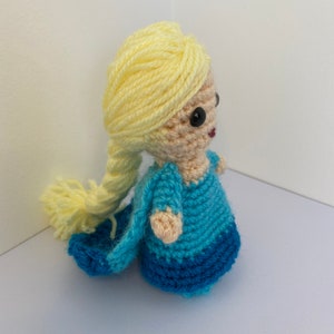 May include: A crocheted doll with long blonde hair, wearing a blue and light blue dress. The doll has black eyes and a pink mouth.