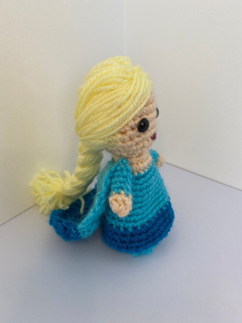 May include: A crocheted doll with long blonde hair, wearing a blue and light blue dress. The doll has black eyes and a pink mouth.