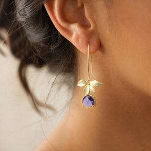 May include: A gold drop earring featuring a long curved wire hook, a cluster of three small matte gold leaves, and a single round faceted purple gemstone dangling at the base. The earring is suspended from an earlobe.