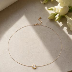 Puede incluir: Un delicado collar de tono dorado con una piedra redonda, transparente y facetada, engastada en un bisel circular dorado. El collar incluye una cadena de hilo fino y flexible y una cadena extensora corta con una pequeña cuenta dorada al final.