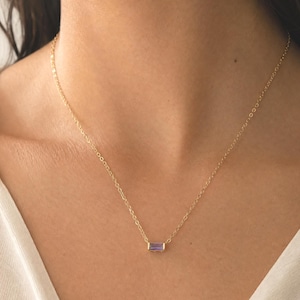 Peut inclure: Un collier à fine chaîne dorée orné d'un unique petit pendentif rectangulaire en pierre précieuse violette taille baguette, serti dans une monture dorée. Le collier repose sur le cou et présente un design à maillons simples avec une finition métallique.