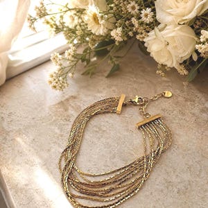 May include: A multi-strand gold-tone bracelet featuring several thin, textured chains connected by two rectangular metal bars at each end. The bracelet includes a lobster claw clasp, an adjustable extender chain, and a small circular metal charm.