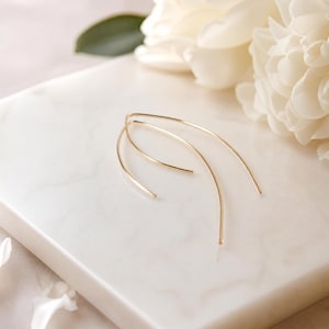 May include: A pair of gold-toned curved wire earrings featuring a slender, elongated arch shape. The earrings are constructed from a single continuous piece of thin metal wire that forms a smooth, open loop design with tapered ends.