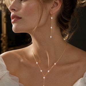 May include: A gold chain necklace featuring small white oval pearls spaced along the delicate links in a Y-shape design, paired with a matching pair of drop earrings, each consisting of a thin gold chain with three small white oval pearls.