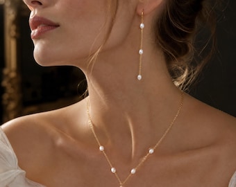 May include: A gold chain necklace featuring small white oval pearls spaced along the chain in a Y-shape design, paired with a matching long drop earring consisting of a thin gold chain and three small white oval pearls hanging vertically.