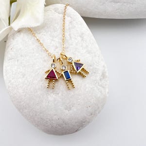 May include: Gold necklace with four charms, each featuring a small figure with a different colored gemstone. The charms are in the shape of a person with a round head and small arms and legs. The gemstones are red, blue, purple, and clear.