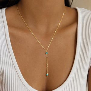 Könnte beinhalten: Eine goldene Halskette mit Y-förmigem Anhänger. Die Kette ist mit zwei kleinen, runden, leuchtend türkisfarbenen Perlen und zwei winzigen goldfarbenen Metallperlen verziert, die entlang des vertikalen Teils der feinen Kette angeordnet sind.