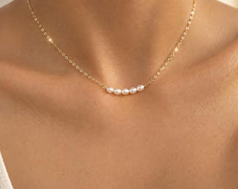 Puede incluir: Un collar de cadena dorada delicada que presenta una hilera de cinco perlas blancas pequeñas y redondas centradas a lo largo de la clavícula. El collar está construido con eslabones dorados finos y pulidos que se conectan a las perlas.