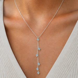 May include: A silver lariat necklace featuring a delicate chain and a vertical drop section adorned with six small, light blue, faceted bicone crystals spaced evenly along the length of the chain, creating a subtle, shimmering, linear design.