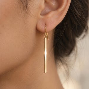 May include: A gold-toned drop earring featuring a long, slender, rectangular bar pendant suspended from a simple curved hook wire. The metal surface has a polished, reflective finish that catches the light, creating a clean and vertical line.