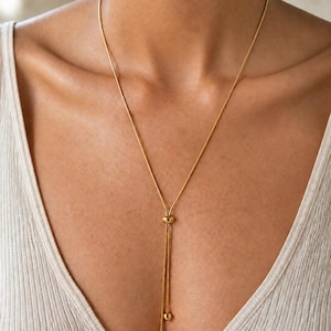 May include: A gold-toned lariat necklace featuring a thin snake chain that passes through a small sliding bead, ending in two dangling strands with spherical gold-toned beads at the tips, resting against a white ribbed sleeveless top.
