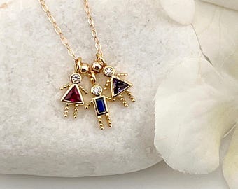 May include: A gold chain necklace featuring three small charms shaped like figures. Each figure has a round clear stone head and a body made of a colored gemstone: a red triangular stone, a blue rectangular stone, and a purple triangular stone.