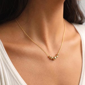 May include: A gold-toned necklace featuring a delicate chain and a central focal point consisting of five polished, spherical beads of varying sizes arranged in a graduated row, resting against the base of the neck.