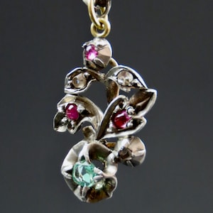 May include: A silver pendant necklace with a floral design. The pendant features red and clear gemstones, along with a green gemstone. The necklace has a delicate chain and a gold-coloured bail.