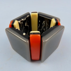 May include: A geometric bracelet featuring gray, black, orange, and cream-colored segments. The bracelet has a unique design with angular shapes and a mix of solid and patterned elements. The bracelet is a bold fashion statement.