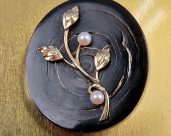Könnte beinhalten: Ovale schwarze Brosche mit goldfarbenem Astdesign mit drei Blättern und zwei Perlen. Die Brosche hat eine Holzmaserung und ist vor einem goldenen Hintergrund platziert. Ein einzigartiges Schmuckstück.