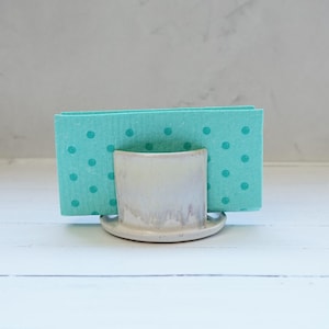 Peut inclure: Un porte-serviettes en céramique beige clair, contenant des serviettes turquoise à pois turquoise. Le support a une base arrondie et une ouverture incurvée pour les serviettes.
