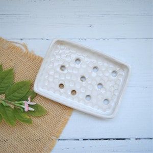 May include: Cream ceramic soap dish with a textured surface and circular drainage holes. The rectangular dish is placed on a burlap cloth with green leaves and pink flowers.