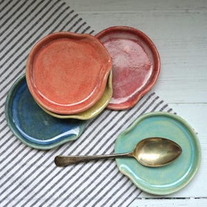 Könnte beinhalten: Eine Sammlung von vier runden Löffelablagen aus Keramik in den Farben Koralle, Dunkelrot, Tiefblau und Hell-Türkis. Jedes Stück hat eine glatte, glasierte Oberfläche und eine kleine Einkerbung am Rand. Ein silberfarbener Metalllöffel liegt auf der türkisfarbenen Ablage.