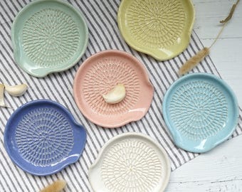 May include: A collection of six ceramic garlic graters in various pastel colors: mint green, yellow, pink, light blue, white, and navy blue. Each grater features a textured surface for grating garlic and a small handle. The graters are arranged on a striped cloth.