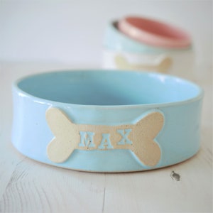 K&ouml;nnte beinhalten: Eine hellblaue Keramik-Sch&uuml;ssel f&uuml;r Haustiere mit einem knochenf&ouml;rmigen Design und dem Namen "MAX" darauf.