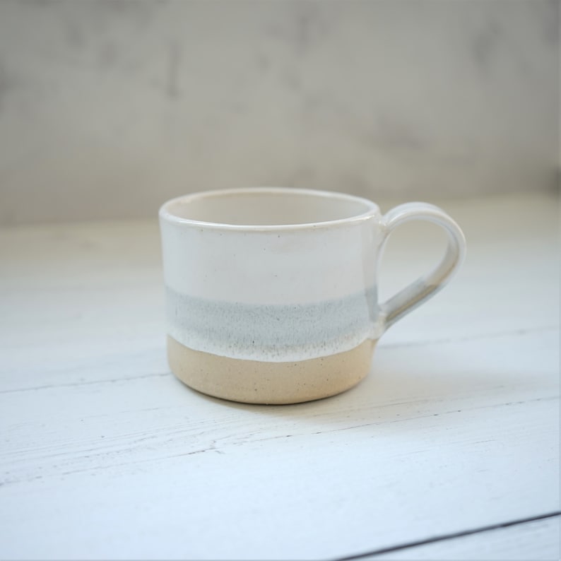 A mug has been glazed in white with a soft grey band running around the mug sitting just below the middle of the mug. The section under the grey band has been left unglazed to show the bare, buff coloured clay.