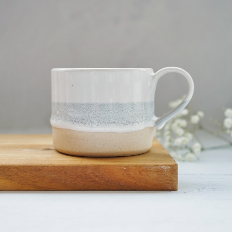 The mug is shown again sitting on a wooden board with a small spray of white flowers in the background.