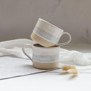 May include: Two ceramic mugs stacked on top of each other, each featuring a three-tone design with a speckled white top, a light grey middle band, and a solid beige base. Each mug has a curved handle on the side and a smooth, rounded rim.