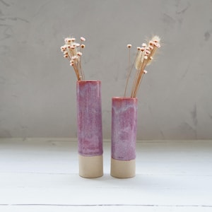 May include: Two cylindrical ceramic vases in shades of pink and beige, each holding dried flowers. The vases are set against a grey backdrop on a white surface. The vases are a decorative home decor item.