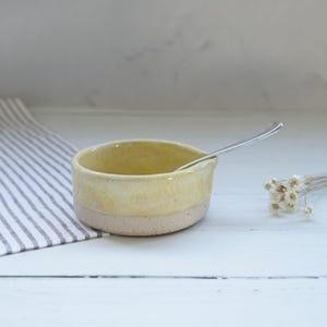 Peut inclure: Un petit bol en céramique fait à la main avec un glaçage jaune clair et une base rose clair mouchetée. Une petite cuillère en argent repose à l'intérieur du bol. Le bol est sur une surface blanche.