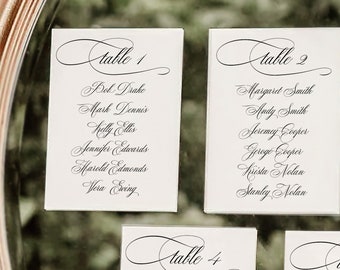 May include: Five white cards with black calligraphy listing table numbers and guest names. The cards are arranged in a row on a mirrored surface with greenery around the edges.