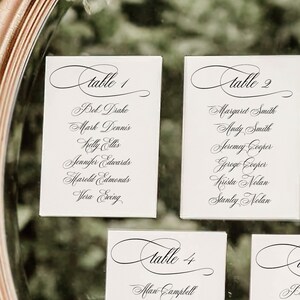 May include: Five white cards with black calligraphy listing table numbers and guest names. The cards are arranged in a row on a mirrored surface with greenery around the edges.