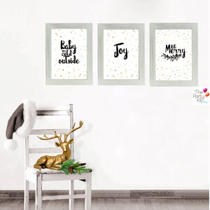 Puede incluir: Tres impresiones enmarcadas con fondos de purpurina dorada y texto negro. Las impresiones dicen "Baby it's cold outside", "Joy" y "Be Merry".