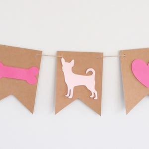 May include: A pink and brown banner with a dog, bone, heart, and dog silhouette. The banner is made of brown paper and has a string running through it.