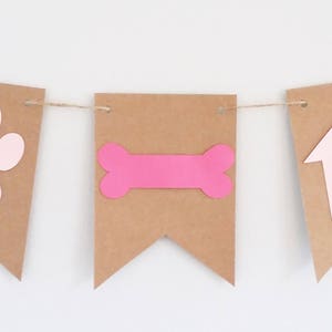 May include: A brown paper banner with pink paw print, bone, and doghouse cutouts. The banner is strung together with twine.