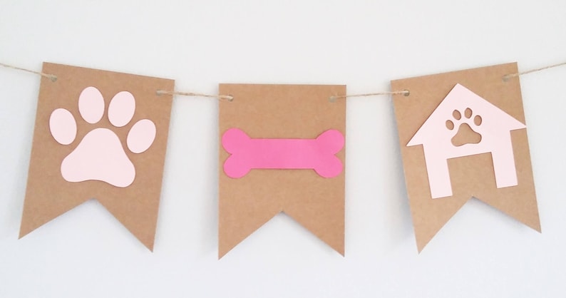 May include: A brown paper banner with pink paw print, bone, and doghouse cutouts. The banner is strung together with twine.