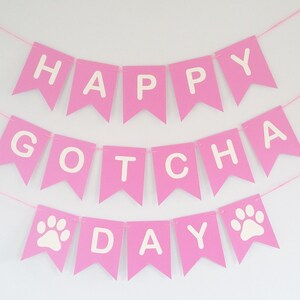 May include: Pink banner with white letters spelling "Happy Gotcha Day" with paw prints on either side of the word "Day".