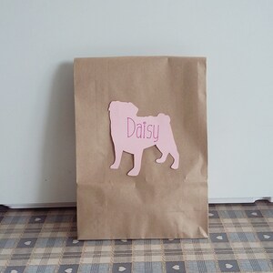 May include: A brown paper bag with a pink paper cutout of a pug dog with the name "Daisy" written on it.