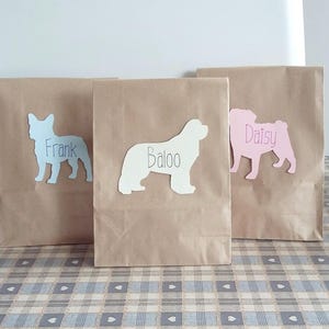 May include: Three brown paper bags with dog silhouettes and names written on them. The bags are on a checkered table cloth. The names are Frank, Baloo, and Daisy.