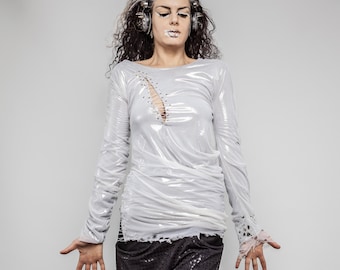 May include: A woman with white makeup and silver headphones wears a white, sheer, long-sleeved shirt with a distressed design. The shirt has a V-neck and is tucked into black pants with a sparkly pattern.