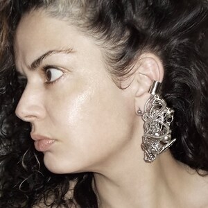 May include: A close-up of a person's face with a large, intricate silver ear cuff. The cuff is made of many small, interconnected pieces and hangs down from the ear.