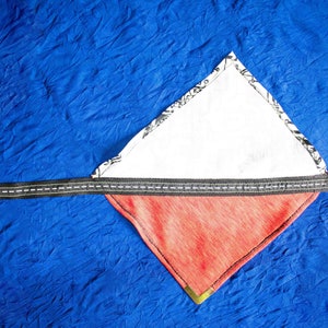Pode incluir: Um bandana vermelho e branco com uma al&ccedil;a preta. O bandana &eacute; dobrado em forma de tri&acirc;ngulo e tem um forro de tecido branco.
