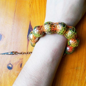 Puede incluir: Un brazalete hecho de una serie de bolas plateadas envueltas en una malla verde y naranja. El brazalete está en la muñeca de una persona.