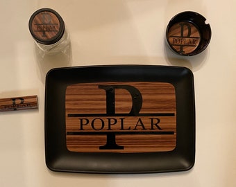 May include: A black tray with a wooden insert featuring the letter "P" and the name "POPLAR" in black lettering. The tray is accompanied by a small black ashtray with a wooden insert featuring the letter "P" and the name "POPLAR" in black lettering, and a small glass jar with a wooden lid featuring the name "POPLAR" in black lettering.