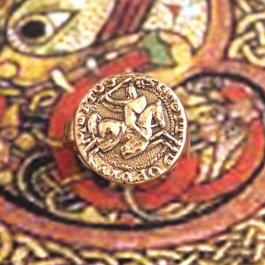 Op de afbeelding: Een gouden ring met een gedetailleerd ontwerp van een persoon die een paard berijdt. De ring is geplaatst tegen een kleurrijke achtergrond.