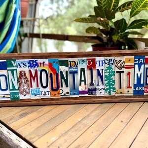 May include: A rectangular wooden sign featuring the text ON MOUNTAIN TIME spelled out using various colorful vintage license plates. The sign includes a graphic of a bear and a pine tree integrated into the letters, all framed in dark wood.