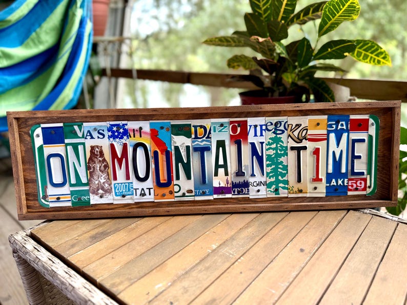 May include: A rectangular wooden sign featuring the text ON MOUNTAIN TIME spelled out using various colorful vintage license plates. The sign includes a graphic of a bear and a pine tree integrated into the letters, all framed in dark wood.