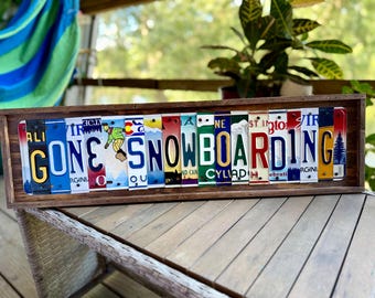 May include: A rectangular wooden frame containing a series of vertical, colorful license plate segments arranged to spell out the phrase GONE SNOWBOARDING, featuring a graphic of a person on a snowboard and various state-themed designs.