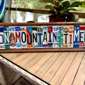 May include: A rectangular wooden sign featuring the text ON MOUNTAIN TIME spelled out using various colorful vintage license plates. The plates display diverse state graphics, including a bear, a pine tree, and various letters, all framed within a dark brown wooden border.